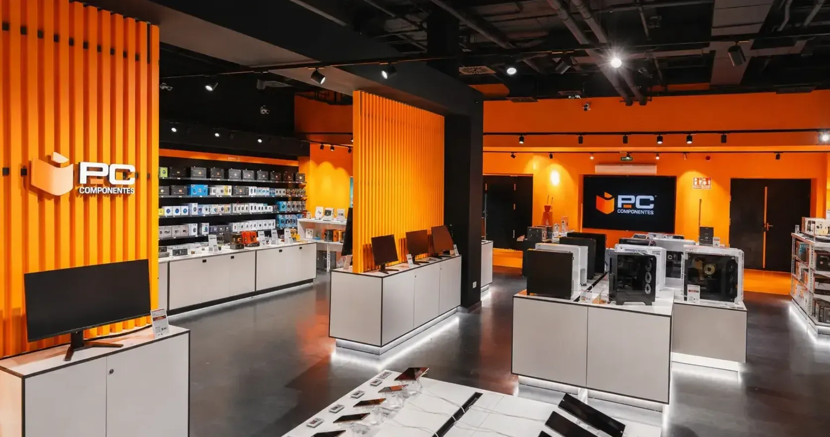 Interior de una tienda física de PcComponentes, con monitores, torres y teléfonos expuestos sobre mostradores blancos entre paneles naranjas